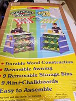 Box showing image of 2-in-1 grocery store and lemonade stand toy with details highlighting durable wood construction, reversible awning, 9 removable storage bins, 9 mini chalkboards, and easy assembly.