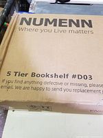Photo of front side of the box showing brand Numenn, 5 Tier Bookshelf label, model #D03, and replaceable parts contact info.