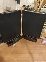 Front view of both speakers upright on wooden floor, showing black cloth grills and walnut-veneer trim.