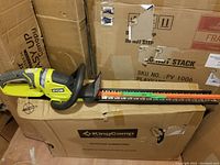 Side view of Ryobi cordless hedge trimmer resting on cardboard boxes. Shows blade guard and front handle.