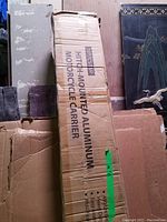 Sealed cardboard box labeled “HITCH-MOUNTED ALUMINUM MOTORCYCLE CARRIER” leaning against wall