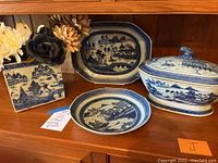 All four pieces arranged on wooden shelf: square vase with flowers, octagonal platter, round shallow dish, and lidded server
