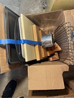 Overall view of boxed items showing stacked metal baking trays, wooden boards, and wire racks held together with a blue strap.