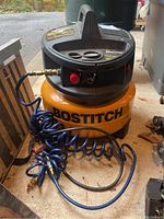 Overall view showing yellow tank with BOSTITCH branding, black top with controls and attached coiled blue air hose