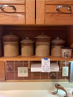 Four ceramic lidded canisters and a ceramic garlic holder arranged in cabinet shelf.