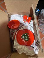 Photo showing red ceramic bowl, small plate, and salt and pepper shakers with Christmas tree motif packed in box with white packing paper