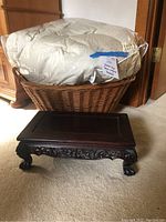 View of queen-sized down comforter in plastic packaging inside large wicker basket, with small dark wood carved footrest in front