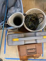 Photo showing Grizzly framing nailer case labeled 'Framing Nail Gun', white bucket with cords, rolled papers, and smaller brown tackle box.