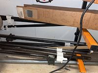 Multiple black metal pipe clamps with white plastic handles resting on a workshop rack, with a cardboard box beside them.