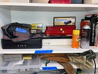 Shelving with the lot showing binoculars, metal lock box, red metal storage boxes, honing stone in box, minimum two spray cans including lubricant oil, and Burris small boxes.