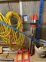 Surveyor's tripod, yellow extension cords hanging, blue drywall T-square, and bucket on floor