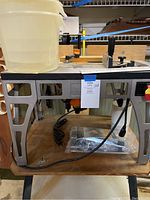 Side view of the additional router table with metal frame and mounted base, wooden top surface, and five stacked clear plastic lidded storage tubs.
