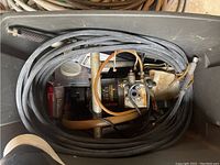 Top down view into a grey plastic tub showing the Wagner airless sprayer with black metal pump housing and coiled black hoses.