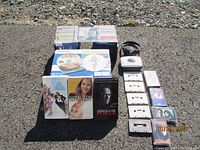 Photo showing the Toshiba Portable DVD Player box with DVDs, Jensen cassette player, cassette tapes, VHS tapes and CDs arranged on pavement