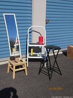 White metal two-step stool with black steps and handle, light wood small step stool with round hole, foldable round black stool, full-length white-framed mirror, outside on asphalt beside storage units.