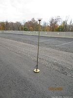 Full view of tall brass torchiere floor lamp standing outdoors on pavement, showing slender pole and circular base.