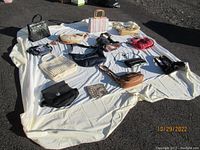 Overview photo of 15 purses laid out on a sheet on an outdoor surface, showing various colors and styles including black, cream, tan, stripe, and floral patterns.
