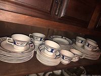 Partial set of Adams Lancaster pattern dinnerware stacked on shelves showing cups, saucers, and plates with blue, green and red leaf pattern on white ceramic.