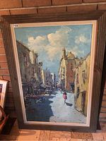 Full view of the large framed oil painting depicting a street scene with people walking, buildings on both sides, under a blue sky with clouds.