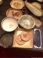 Overall view of pottery pieces: handled casseroles with botanical and rooster motifs, fish-shaped vase, small red/black vase, various bowls, platters, trays and plastic container