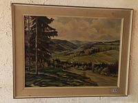 Framed landscape painting showing a rural valley with trees and hills under a cloudy sky.