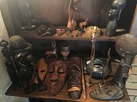 Overall display of two shelves showing multiple carved wood masks and sculptures