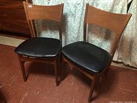 Front view showing two mid century modern wooden chairs with black cushioned seats and slightly curved wood backrests, placed on a brown floor with curtains in background.