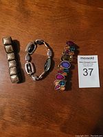Top-down photo of three bracelets laid side by side on wooden surface—multi-color stone set bracelet on right, brass-brown bracelet on left, black-silver-gold linked bracelet in middle