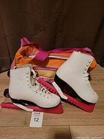 White CCM ice skates placed on brown surface, pink blade guards attached, showing skate sides and laces