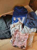 Full view of clothing lot showing sweatshirts, t-shirts, and pants arranged on floor near wall.