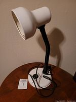 White desk lamp with flexible black neck and round base shown from side angle with cord coiled on base. The lamp is placed on a wooden surface against a beige wall.