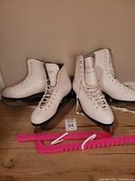 Two pairs of white figure skates with visible laces and high ankle support. Pink blade protectors placed in front of the skates on the floor.
