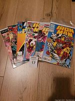 Six Iron Man comic books arranged side by side on wooden floor, with themes showing various Iron Man cover arts and a white maxsold lot number tag.