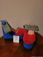 Side and front view of the shoes showing red suede upper, green ankle strap with blue button, and blue/black platform wedge heel.