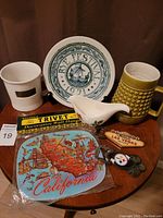 Vintage kitchen collectibles displayed on wooden table: Rye Festival 1971 plate, California decorative trivet, floral ceramic pitcher, green textured mug, Las Vegas and Steelers keychains.