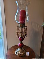 Full view of the candle holder showing the curved clear glass chimney containing a red candle inside, the brass and red spherical base, and hanging decorative beads.