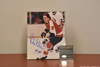 Signed 8x10 color photo of Bill Barber on ice in Flyers uniform with blue ink signature and HOF 1990 inscription, displayed upright on wooden surface with Certificate of Authenticity card next to it.