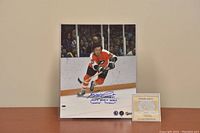 Photo showing the signed 8x10 action photo of Reggie Leach skating in Flyers orange and black uniform with blue ink autograph and inscriptions. Certificate of Authenticity displayed in front.