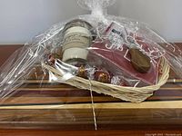 Charcuterie board gift basket wrapped in clear cellophane showing the board, candle, towels, and cheese knives inside a basket with decorative Lindt chocolates.
