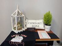 View of Small Town Christmas black sweatshirt laid out with other items on table showing pillow, lantern, planter, and journals.
