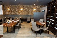 Interior view of the restaurant showing modern wooden tables, upholstered chairs, decorative wood log wall, and wine rack.