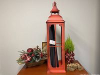 Photo of a red metal lantern on a table with a rustic floral centerpiece, a fresh lemon cypress plant wrapped in plaid, and a cheese board set with cheese knives in a box.
