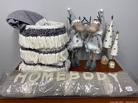 Photo shows a grey Home Body pillow, a grey and cream woven storage basket, two silver moose figurines dressed in winter clothing, and two white Christmas tree decorations with gold bell accents.
