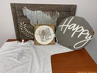 Photo showing the wall decor, round grey pillow with 'Happy' text, glass plate set with napkins and spreader, and grey sweatshirt laid out on a wooden table.