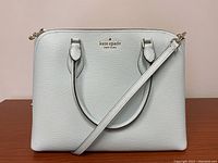 Front view showing teal Saffiano leather and gold-tone logo, handles, and crossbody strap placement