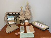 Overview of wellness gift kit items arranged on wood table showing beige knit hat, vegan suede pencil case, journals, candle, small emergency and travel kits, faux fur coin purse, scarf, and two Christmas tree decorations.