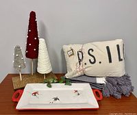 Photo shows the entire set of five items: ski-themed serving platter with red handles, three cone-shaped yarn Christmas trees in red, white, and grey with white pom-poms, a folded two-tone throw blanket, and a cream-colored pillow with embroidery.