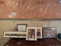 Lot of five art pieces displayed on a table with visible metal easels and protective cellophane on prints.