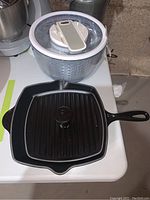 Le Creuset cast iron square griddle pan with ridged surface and press alongside Zyliss salad spinner on white surface.