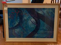 Framed multimedia abstract painting displayed upright on a wooden floor with a mostly blue and purple swirling organic design and red marks near top left.